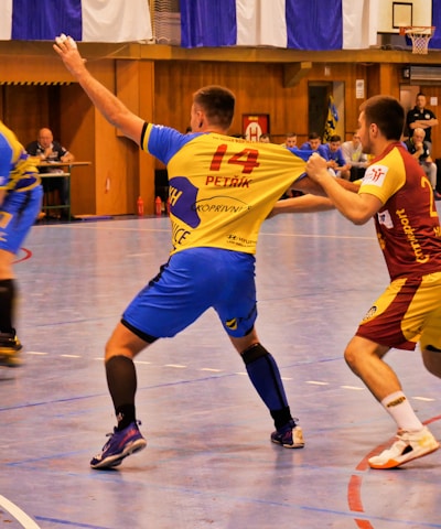 A dynamic indoor sports scene with players in motion on a court. Two athletes in blue and yellow uniforms are engaged in an intense handball game, with one player gripping the other's jersey. The background shows other players, several seated people, and advertisements on the walls.