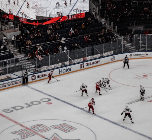 A professional ice hockey game is in progress inside an arena, with players in red and white jerseys actively engaging on the rink. The scoreboard above displays the score and game details, while spectators are seated in the stands. A large screen shows a live feed of the match action.